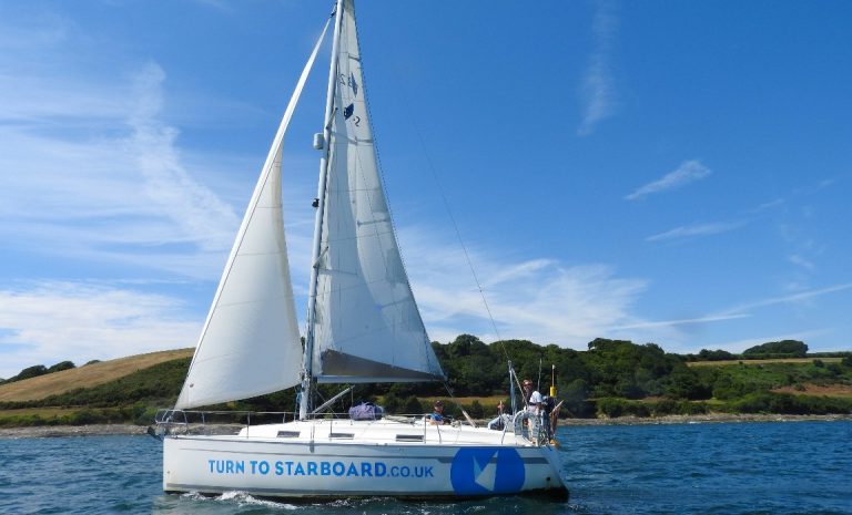 A white sailboat on the tranquil ocean, with "TURN TO STARBOARD.CO.UK" emblazoned on its side, sails gracefully near a hilly coastline under a clear blue sky. The vessel, often crewed by ex-soldiers exploring new career horizons, embodies serenity and adventure.