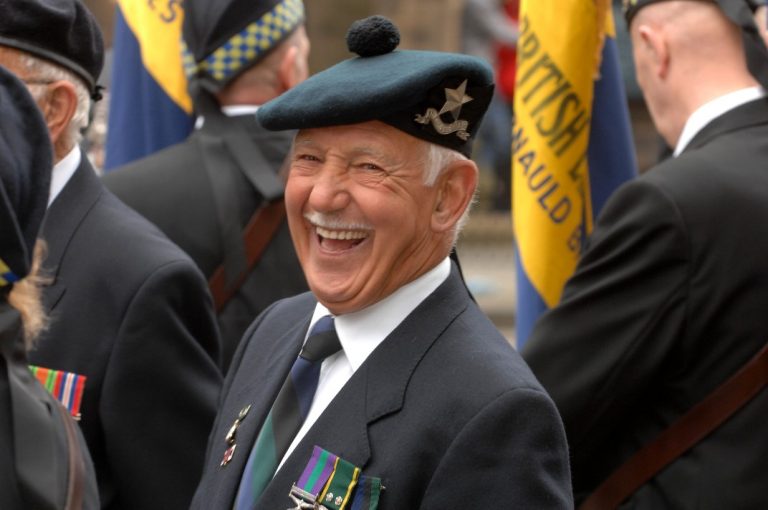 An elderly man in military uniform grins warmly, proudly displaying his medals. Standing among fellow veterans with vibrant banners in the background, he embodies the spirit of camaraderie and honor at this Veterans Project, a ground-breaking initiative celebrating heroism.