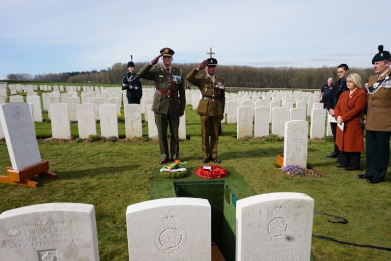 Soldiers saluting at a military cemetery, with white gravestones marking the resting place of unknown soldiers buried over 100 years ago, while a group of civilians and a bagpiper stand nearby.
