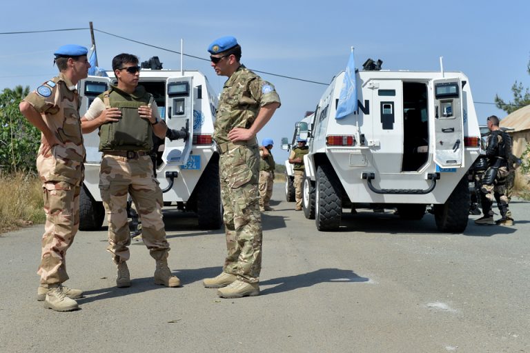 UN peacekeepers in uniform stand near white armored vehicles on a rural road in Cyprus, engaging in discussion about strengthening defence ties.