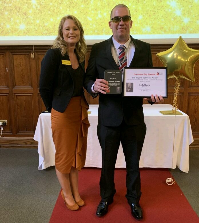 A man in a suit and sunglasses, who is a blind veteran, proudly holds a certificate and plaque. He stands alongside a woman in business attire before a gold backdrop, beside a table with a gold star balloon. They've just received the highest honor from the charity organization they're part of.