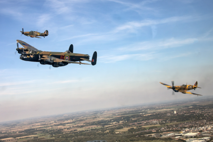 Three vintage military aircraft, a tribute to the brave veterans of D-Day, soar in formation over a landscape with fields and distant towns visible below.