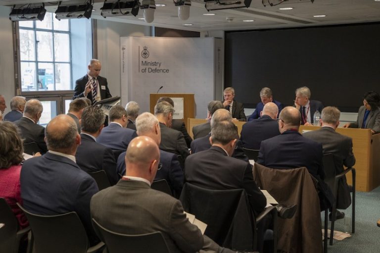 A meeting in progress at the Ministry of Defence highlights collaboration, with attendees seated in rows facing a presentation area. The Minister stands as the speaker, while others are seated at a panel table.