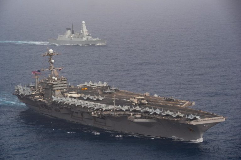 A US aircraft carrier and a Type 45 destroyer navigate side by side on the ocean near Malta, with the carrier's deck lined with aircraft.