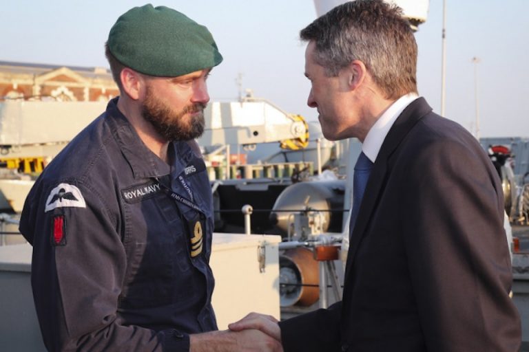 Two men, one in a military uniform with a beret, the other in a suit, shake hands on a ship's deck, symbolizing an investment poised to revolutionize naval operations.