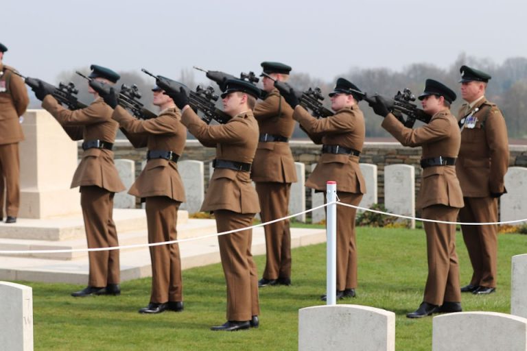 Seven soldiers in brown uniforms perform a solemn rifle salute at a cemetery, honoring those laid to rest from World War 1.