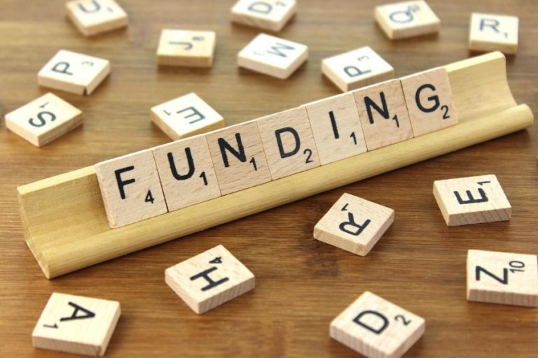Wooden Scrabble tiles spell "FUNDING" on a tile rack, hinting at a grant awarded, surrounded by random tiles on a wooden surface.