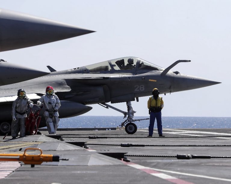 A fighter jet on an aircraft carrier deck, ready for counter-Daesh operations, is surrounded by three crew members in protective gear and another person in blue standing nearby. The scene unfolds with precision akin to a Duncan & French production.
