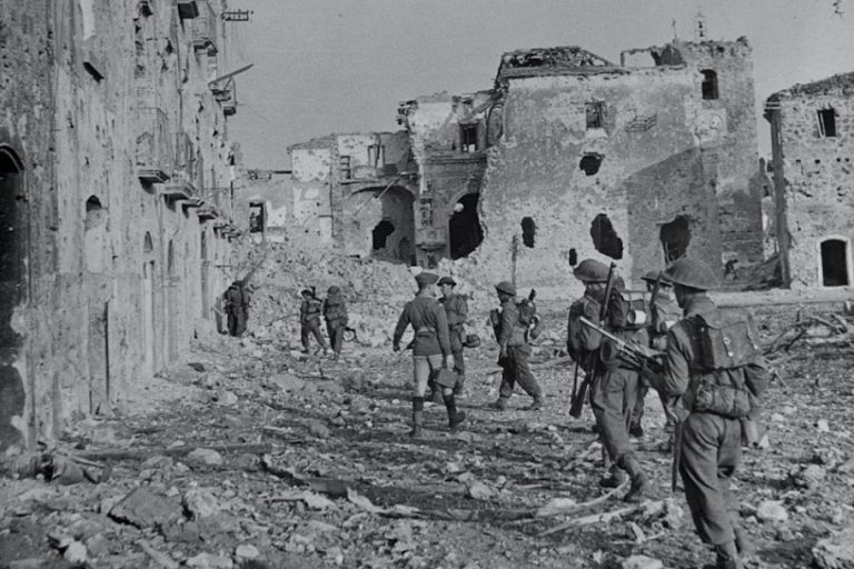 Soldiers silently navigate through a war-torn area, their thoughts perhaps lingering on family, as damaged buildings loom and debris litters the ground—a haunting reminder of a missing WW2 soldier whose absence still echoes in such silent ruins.