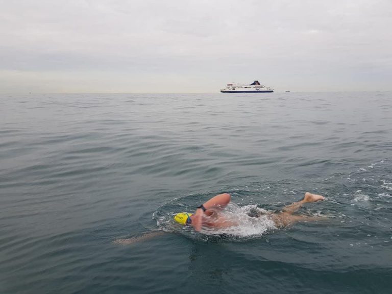 In 2019, a swimmer with a yellow cap bravely takes on The Channel, embodying the spirit of the Victory Swim. A ship looms in the distance under a canopy of clouds.
