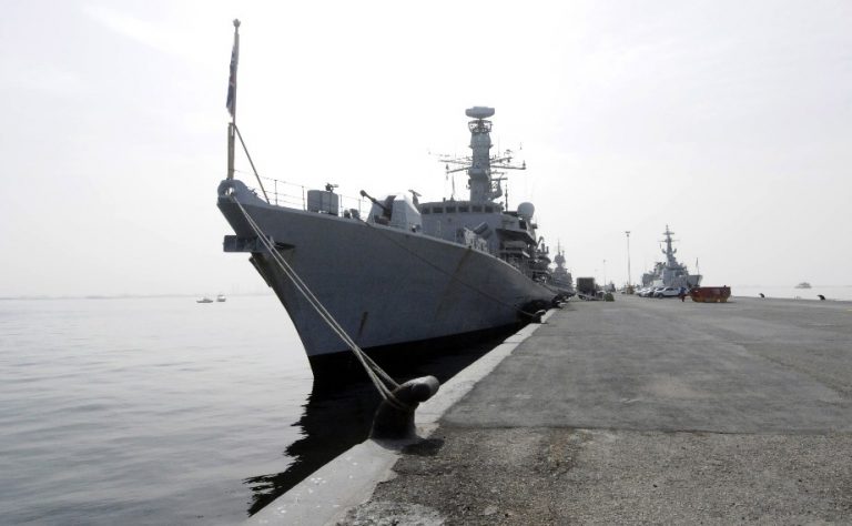 Military ships docked at a harbor for the UK's Gulf Mission, with one ship prominently in the foreground and another in the distance under a cloudy sky, marking a new era of strategic presence.