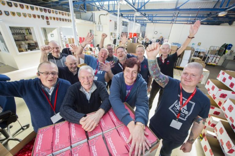 A group of eleven people in a refurbished warehouse environment, smiling and raising their hands, is surrounded by boxes with poppy designs at the historic Poppy Factory.