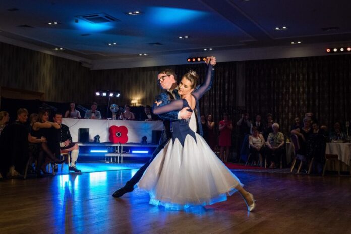 The couple, who raised £25k for charity, performs a ballroom dance on the polished wooden floor, with an audience seated in the background. The room is dimly lit with blue accent lights as they danced gracefully across the stage.