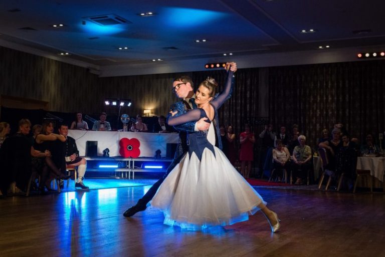 The couple, who raised £25k for charity, performs a ballroom dance on the polished wooden floor, with an audience seated in the background. The room is dimly lit with blue accent lights as they danced gracefully across the stage.