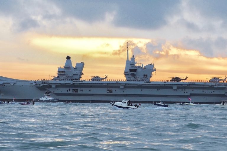 An aircraft carrier sails under a cloudy sky, with helicopters poised on deck and small boats flanking its sides, symbolizing MOD testing as part of a billion-pound investment in maritime excellence.