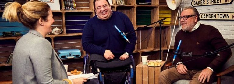 Three people are sitting in a room with microphones, smiling and engaging in conversation about resilience. Shelves with books and decor adorn the background, adding to the cozy atmosphere.