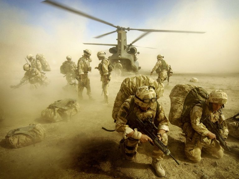 Marines in tactical gear disembark from a helicopter, crouching and securing the area with weapons ready, amidst a dusty environment.