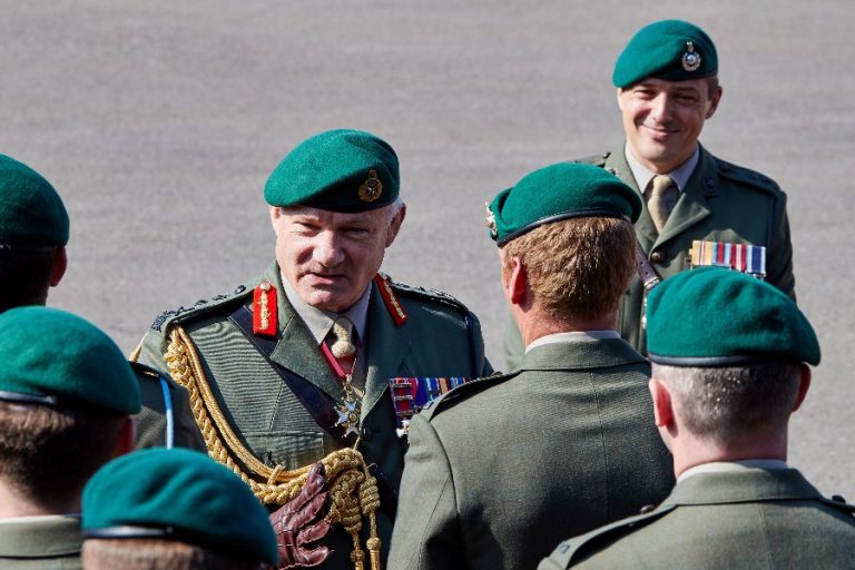 A group of uniformed Marines, wearing green berets, gather outdoors. The central figure appears to be an officer with decorated medals and an ornate sash, reminiscent of a Peace Prize winner's honor, speaking to other members.