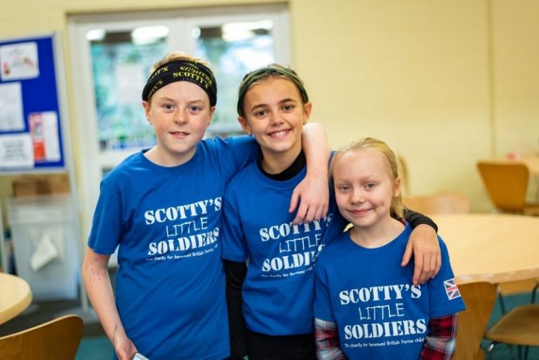 Three children wearing matching "Scotty's Little Soldiers" t-shirts stand together in a room, smiling at the camera, as they gear up for the exciting Marathon Returns this month of May.