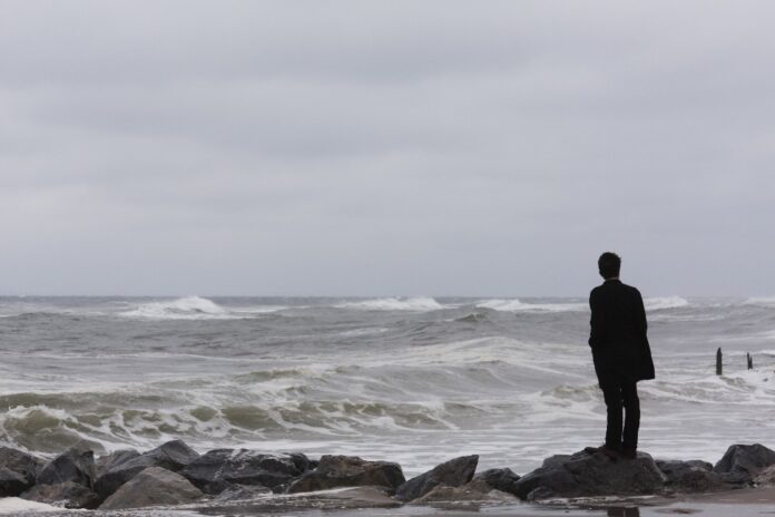 A lone character in a dark coat stands on the rocks by a stormy ocean, gazing at the waves under a cloudy sky, as if they are about to become part of some unfolding thriller.