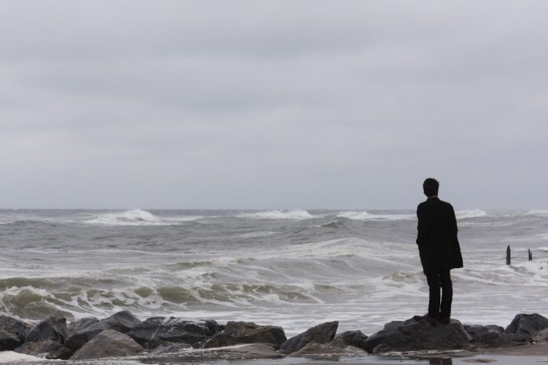 A lone character in a dark coat stands on the rocks by a stormy ocean, gazing at the waves under a cloudy sky, as if they are about to become part of some unfolding thriller.
