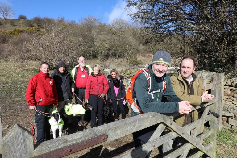 A group of people with dogs stands by a wooden gate in a rural area, facing life's challenges together, with trees and a hill in the background.