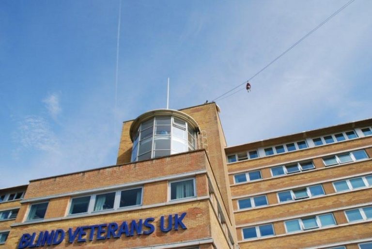 A person is taking on the Zip Wire Challenge, soaring off the roof of a brick building adorned with "Blind Veterans UK" signage. The sky is clear and blue, adding to this thrilling test of mettle.