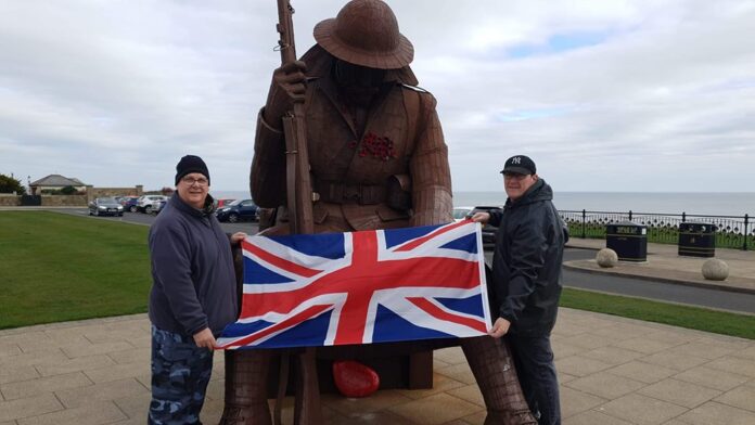 Two people hold a Union Jack flag in front of a seated Tommy statue, with poppies on the chest, near a coastal setting.