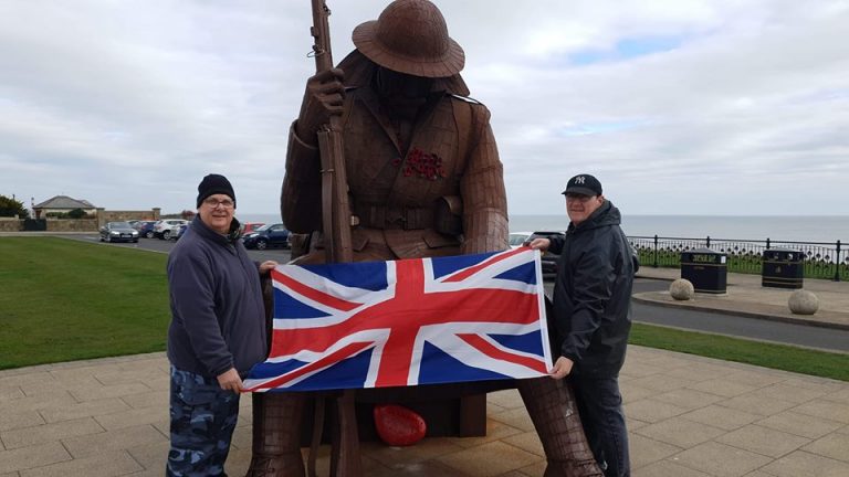 Two people hold a Union Jack flag in front of a seated Tommy statue, with poppies on the chest, near a coastal setting.