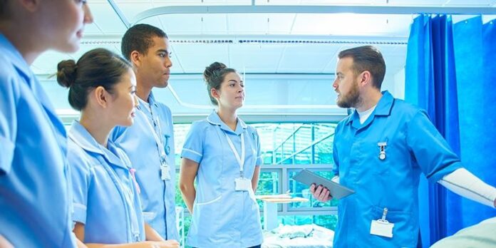 A group of medical professionals in blue uniforms stands in a hospital room, engaging in conversation about the Erskine Veterans Fund. One holds a tablet, detailing how they can provide support to those who served.