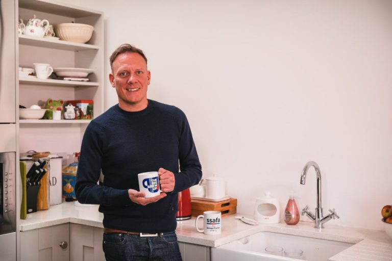 Antony Cotton is standing in a kitchen, holding a mug as he brews up his favorite drink. In the background, shelves lined with dishes and the sink reflect his warm connection to the forces community.
