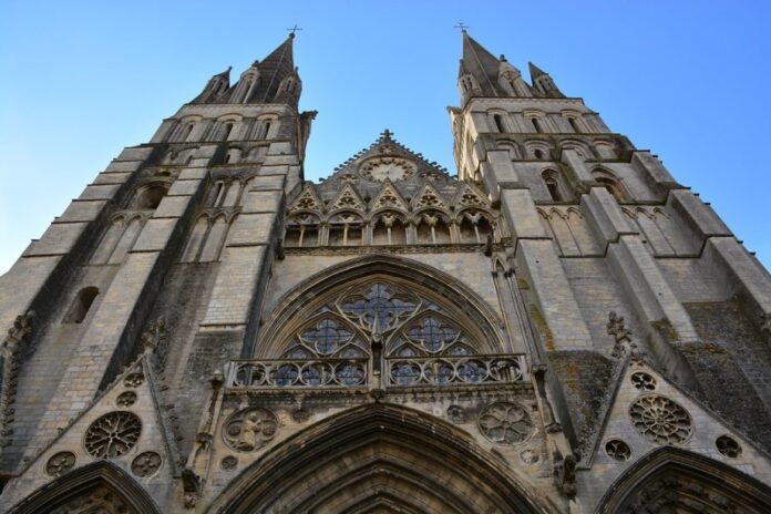 Low-angle view of a Gothic cathedral facade with tall spires, intricate stonework, and pointed arches reaching into the clear blue sky, reminiscent of the solemn beauty found in Normandy's historical architecture.
