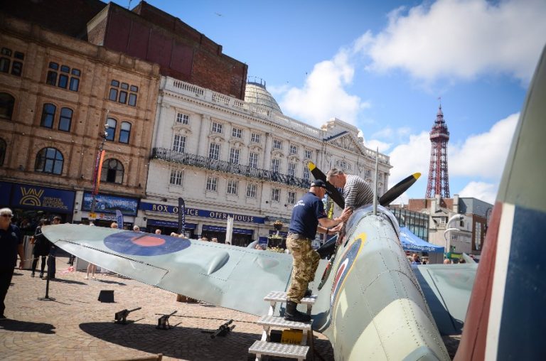 A man inspects a historic fighter plane on display in a city square during Armed Forces Week 2019, with Blackpool's buildings and tower silhouetted against the partly cloudy sky.
