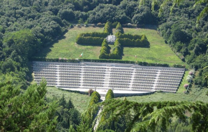 An aerial view of the terraced cemetery at Monte Cassino, where verdant greenery embraces rows of stone graves, memorializing those who fought bravely. A cross-shaped arrangement of bushes in the background serves as a solemn tribute to the departed souls.