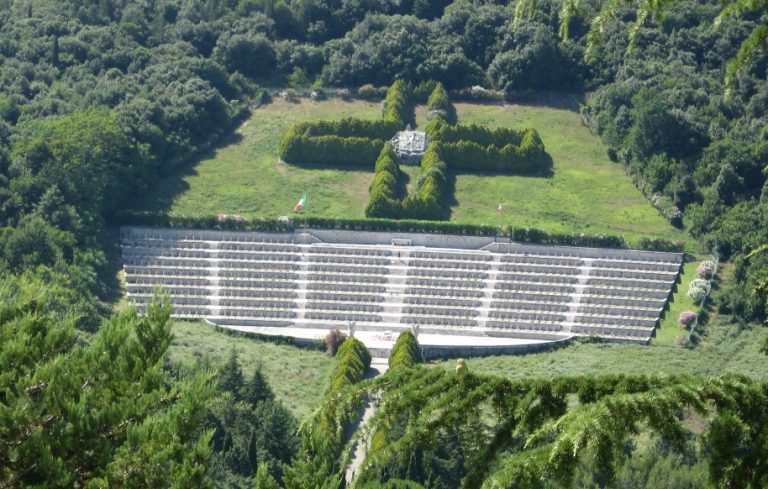An aerial view of the terraced cemetery at Monte Cassino, where verdant greenery embraces rows of stone graves, memorializing those who fought bravely. A cross-shaped arrangement of bushes in the background serves as a solemn tribute to the departed souls.