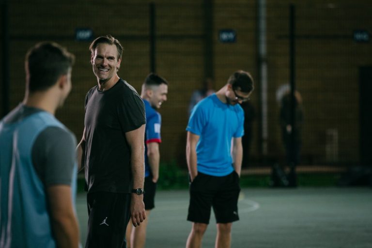 Several men, including the Prince of Wales, stand on an outdoor sports court, some smiling and talking in their casual sports attire.
