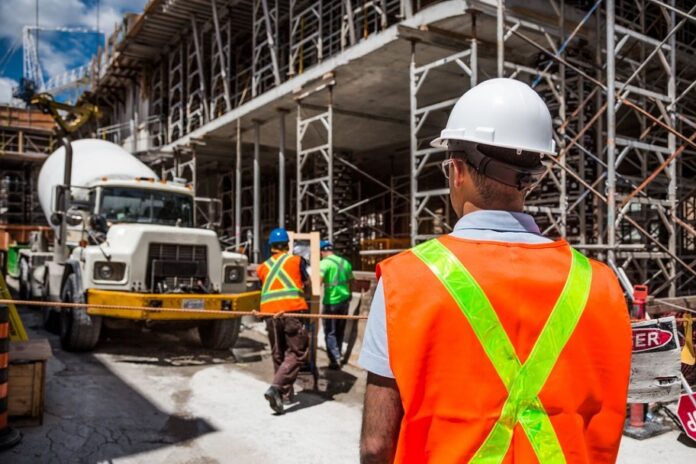 A bustling construction site features a cement truck and workers in hard hats and safety vests maneuvering around the scaffolding, all contributing to a significant building milestone achievement.