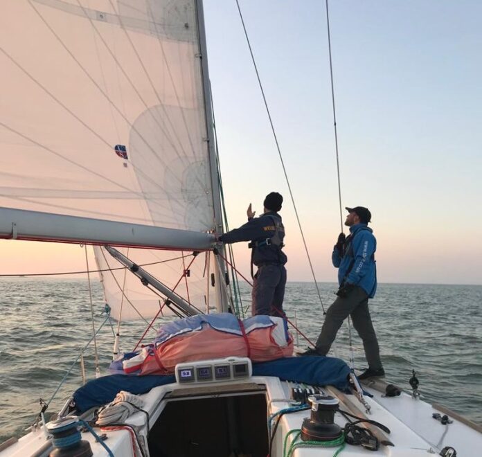 As the sun dips below the horizon, two people expertly adjust the sails on their small sailboat, preparing for a thrilling Cowes-Cherbourg race. The golden glow casts a warm hue over their teamwork at sea, embodying the spirit of camaraderie and charity.