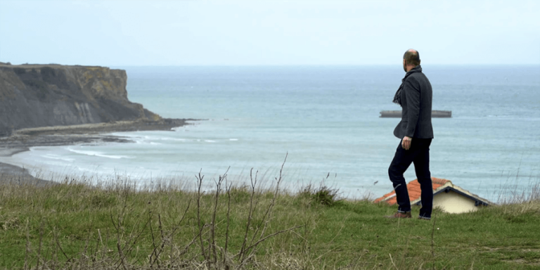 A person stands on the grassy expanse near the coast of Normandy, gazing at the ocean with majestic cliffs in the distance and a quaint house nearby.