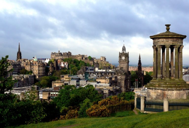 View of the Edinburgh skyline from Calton Hill, featuring historic buildings and lush greenery. The iconic Dugald Stewart Monument stands proudly in the foreground under a cloudy Scottish sky, evoking a sense of history and honoring veterans who have shaped Scotland's legacy.