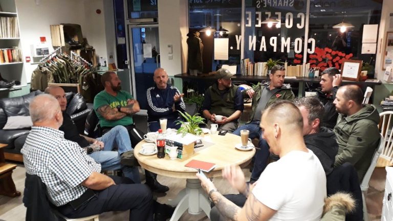 A group of veterans sits in a coffee shop, engaged in conversation around a table with drinks and books, sharing stories and camaraderie.