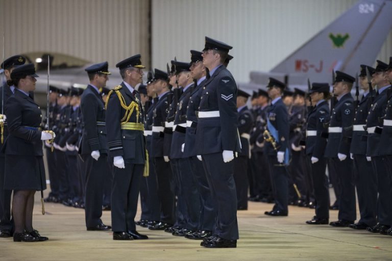 A formal military inspection with uniformed officers in formation inside a hangar at Lossiemouth, conducted by a senior officer scrutinizing the squadron. An aircraft tail looms in the background, ready for Quick Reaction Alert.