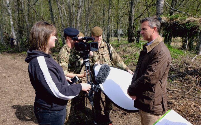 In an Estonian forest setting, a woman interviews a man with a camera crew in military uniforms, capturing the activities of UK troops as they hold their equipment.