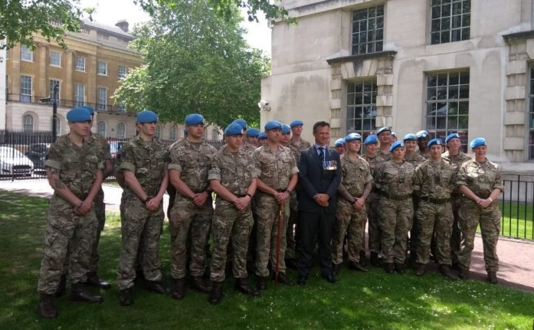 A group of uniformed military personnel, including servicewomen, and a man in a suit pose for a photo outdoors near a building and a lawn, under the shade of trees—embodying the commitment to peacekeeping efforts.