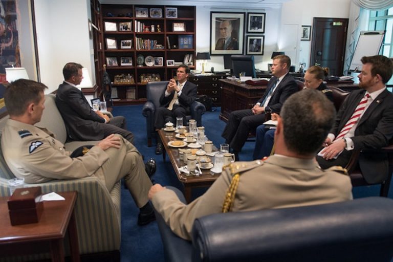 Six people in military and formal attire, including a minister from Washington, are seated around a coffee table in an office engaged in discussion. Drinks and snacks adorn the table, while shelves with books and a large portrait provide the backdrop.