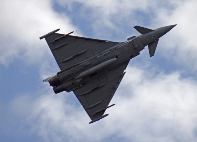 A gray fighter jet, equipped with miniature missile decoys, flies through the cloudy sky, viewed from below at an angle.