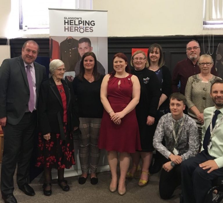 A group of veterans and supporters pose proudly indoors beneath a "Glasgow's Helping Heroes" banner, as part of a minister visit celebrating their dedication and service.