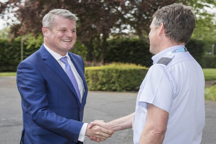 Two men shaking hands outdoors amidst lush trees and bushes, one wearing a blue suit and tie, the other in a light blue shirt with epaulettes, reminiscent of uniforms worn by US pilots after a simulation exercise.