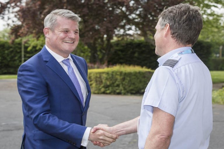 Two men shaking hands outdoors amidst lush trees and bushes, one wearing a blue suit and tie, the other in a light blue shirt with epaulettes, reminiscent of uniforms worn by US pilots after a simulation exercise.