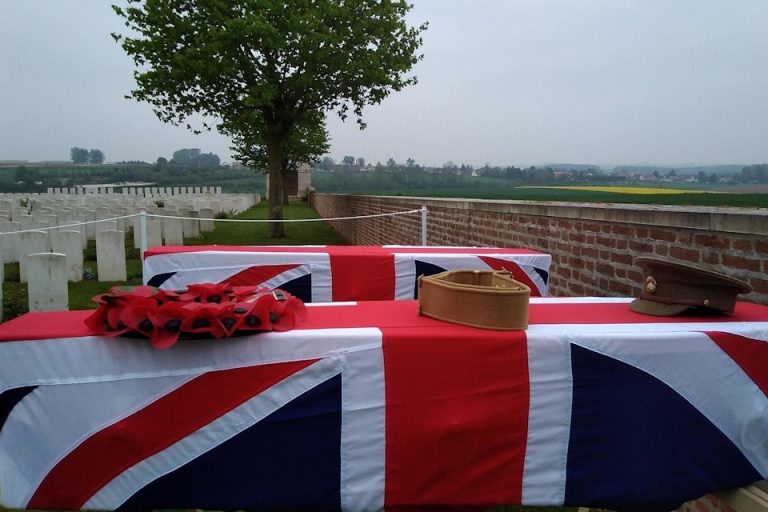 Two coffins draped with Union Jack flags rest outdoors, each adorned with a military hat and poppy wreath, signifying a solemn military burial. They stand in a cemetery against the backdrop of a tree and rows of headstones, honoring soldiers who bravely fought in the Great War.